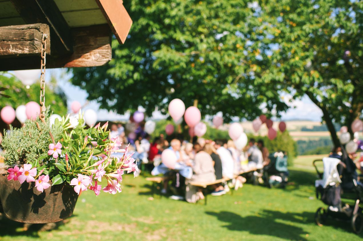 Fröhliche Bauernhof-Feier im Freien mit Gästen an langen Tischen, Ballondekoration und blühenden Blumen unter schattigen Bäumen.